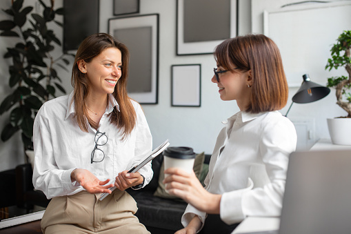 Cheerful lady coworkers enjoying coffee break in office, two businesswomen talking, woman holding takeaway cup, meeting and chatting in office