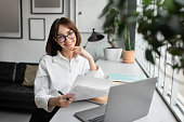 Easy paperwork. Happy female financial advisor using laptop and holding papers, working in cozy office and smiling