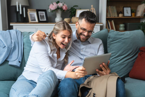 istock Excited young couple sitting on couch in formal wear with tablet pc, celebrating online win, great deal or business success at home. Millennial spouses enjoying big sale in web store. 1864701708