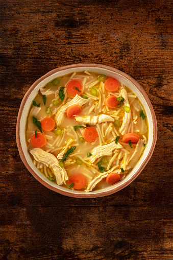 Chicken noodle soup with vegetables, a bowl of healthy broth, overhead flat lay shot on a rustic wooden table, winter comfort food with copy space