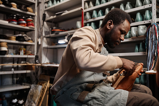 Young African American male owner of shoemaking workshop fixing upper part of unfinished boot to sole while sitting by workplace