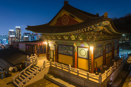The ornate eaves of the Daeseongsa Temple high on Mt. Umyeonsan illuminated at night overlooking the glittering skyscrapers of Seoul’s crowded cityscape, South Korea.