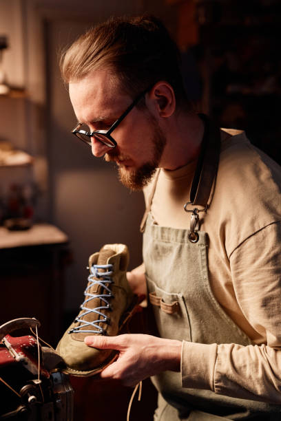 istock Side view of young craftsman in eyeglasses and apron stitching boot workpiece 1864806303