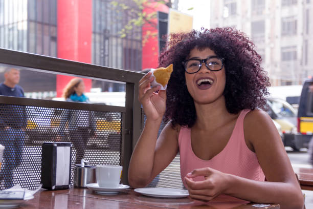 Pretty young woman with afro hairstyle sitting outdoors drinking a cup of coffee and eating coxinha food. Brazilian snack in Sao Paulo during summer, busy street.