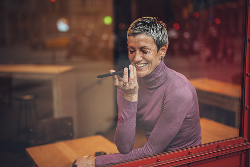 A cheerful mature woman enjoys a lighthearted conversation on her phone, set against the warm glow of a café's interior.