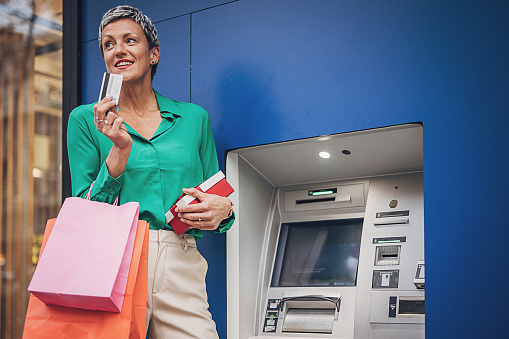 A senior woman with a credit card at an ATM, holding shopping bags and a gift, against a vivid blue background, balancing spending with financial management.