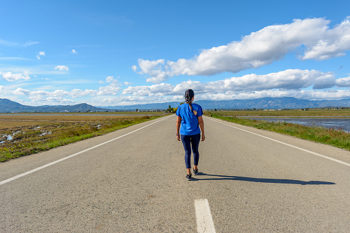 A person is walking down an open road surrounded by nature under a cloud-streaked blue sky, back view of latina woman dressed in blue walking down the road in the Ebro Delta natural park, Tarragona, Catalonia, Spain,