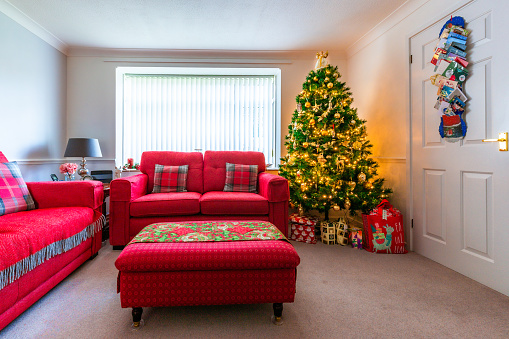 Interior of a domestic living room decorated for the Christmas holidays. The red sofas and green Christmas tree add to the festive feeling. The tree is illuminated and adorned with decorations, and presents underneath the tree.