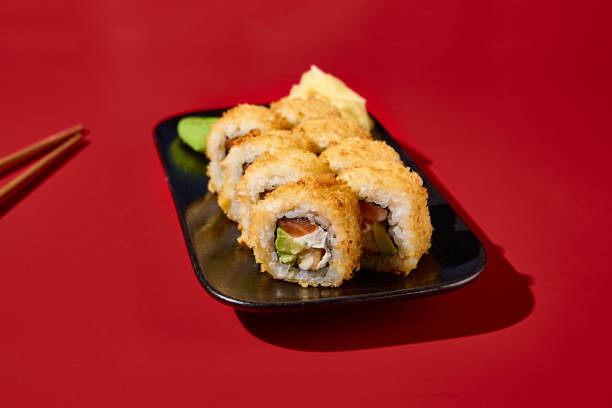 nahaufnahme einer heißen tempura-sushi-rolle mit lachs und avocado, elegant auf einem schwarzen teller mit stäbchen platziert. tiefroter hintergrund intensiviert die atmosphäre der speisekarte eines asiatischen restaurants - 16611 stock-fotos und bilder