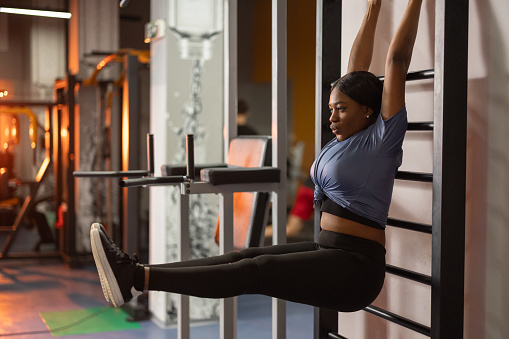 Beautiful black woman hangs on the wall bars and raises her legs in the gym