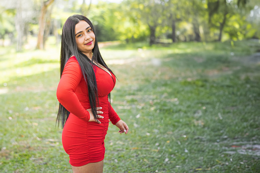 young latina female student, standing in the middle of an outdoor park posing with her hand on her waist.