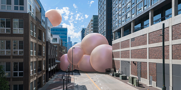 A group of huge pink spheres stuck together on a downtown city street in-between tall buildings. One sphere is floating above the buildings, on a bright sunny day with light clouds.
