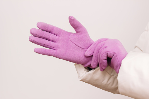 A cropped shot of a woman in a warm jacket putting purple winter gloves on her hands isolated on a light background. Winter clothes