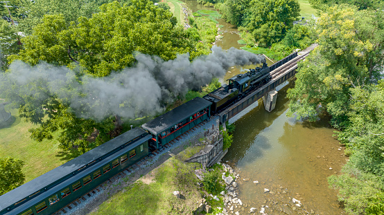 Steam train with smoke from the locomotive driving in the countryside with smoke coming from the chimney. The black and red locomotive is pulling passenger railroad car with tourists.