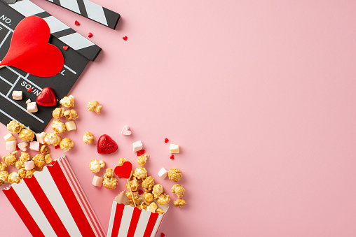 Romance on screen this Valentine's. Top view featuring clapperboard, striped popcorn containers, heart-shaped ornaments, candies, marshmallows, sprinkles on sweet pastel pink surface with advert space