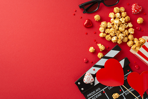 Love Reel Unveiled: Overhead view capturing a clapperboard, 3D glasses, scattered popcorn, chocolates, and heart-shaped decorations on a red surface, announcing a romantic film premiere