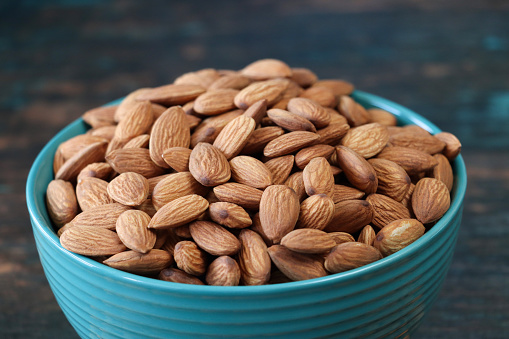 Stock photo showing close-up view of some shelled almonds that are piled high in a turquoise dish, against a blue woodgrain background. Raw almonds are considered to be a very healthy snack food, containing vitamin E, antioxidants and protein, and boasting a list of health benefits while aiding blood sugar control.