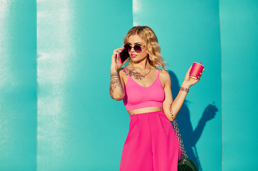 Sunny style fashion woman on blue background. Girl wearing cute trendy pink outfit, drinking tasty coffee, enjoy her walk. Fashion woman doll in glasses, pink clothes hold phone.