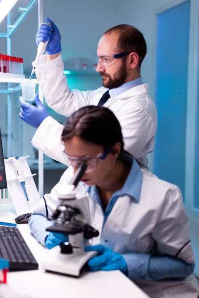 istock Scientist taking sample with micropipette from test tube and asistant analyzing virus on blood through microscop 1865338350