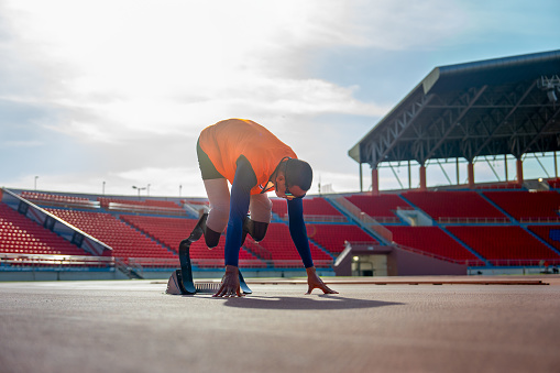 Sport man athlete prosthesis legs with orange cloth stay in the position of start running and look forward at the stadium with soft light of evening.