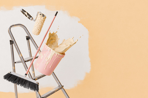 3D rendering of a Paint roller and brush in mid-air with a bucket tipping over on a ladder, spilling paint, against a half-painted wall with copy space.
