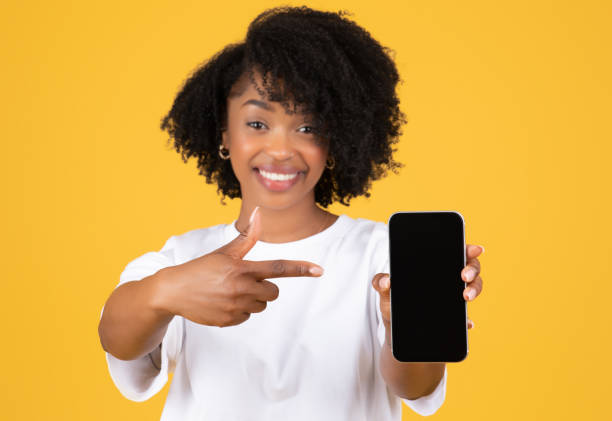 istock Glad millennial black lady in casual pointing finger at phone with empty screen 1865382387