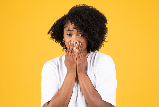 Sad shocked millennial african american curly woman in white t-shirt presses hands to face, crying, isolated on yellow studio background. Tears of joy, good or bad news, ad and offer, emotions