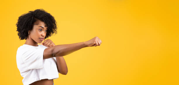 Serious confident millennial african american curly woman in white t-shirt fighting, punching Serious confident millennial african american curly woman in white t-shirt fighting, punching, isolated on yellow studio background, panorama. Self-defense, battle, strength, power, ad and offer self defense stock pictures, royalty-free photos & images