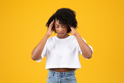 Despaired tired millennial african american curly woman in white t-shirt presses hands to temples, suffering from headache, isolated on yellow studio background. Health problems, fatigue, migraine