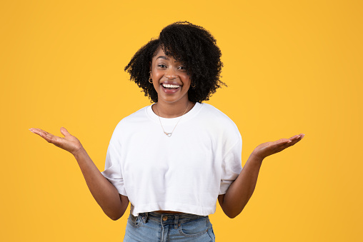Happy surprised young black curly woman in white t-shirt spread arms to sides, hold empty space, isolated on orange background, studio. Choose, lifestyle, ad and offer, human emotions