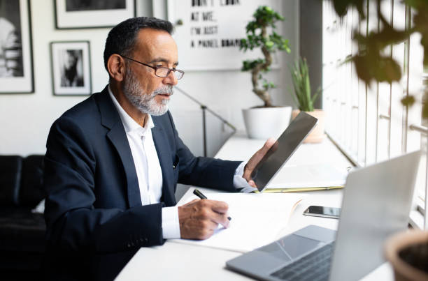 istock Confident serious mature european man in suit work on laptop, makes notes, use tablet for checking account 1865408425