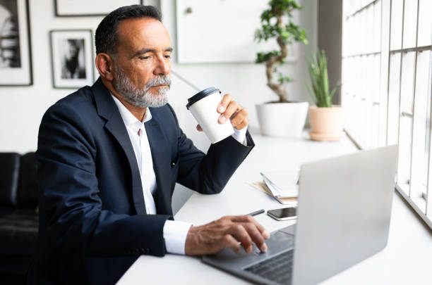 istock Serious mature european man in suit work on computer, drink coffee takeaway, planning project 1865408453
