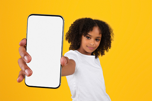 Cheerful teen curly black girl in white t-shirt show smartphone with blank screen, recommends app, ad, offer, isolated on yellow studio background. Blog and website for education, knowledge study
