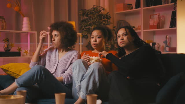 Three women friends eat popcorn while watching a horror movie on couch at home