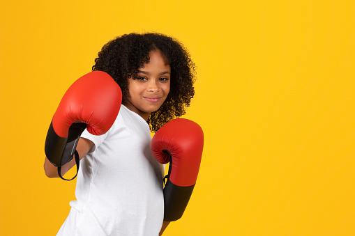 Cheerful strong curly teenage black girl in white t-shirt and red boxing gloves fights, beats isolated on yellow background, studio. Victory, competition, sport and study, sport, ad and offer