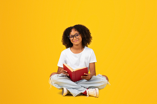 Cheerful smart curly teenager black schoolgirl in white t-shirt and glasses reads book, isolated on yellow background, studio. Study, knowledge, hobbies, education and homework, exam preparation