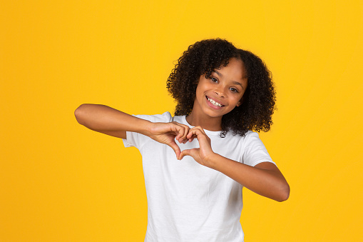 Positive curly teen black schoolgirl in white t-shirt has fun, making heart gesture with hands, isolated on yellow background, studio. Lifestyle, study, knowledge, gratitude and love sign, offer