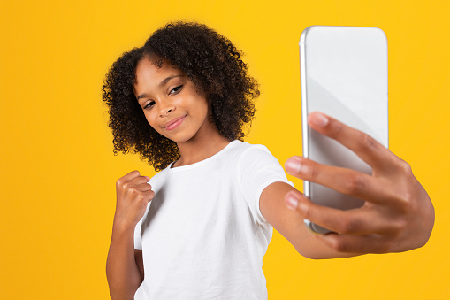 Strong glad teenage black pupil girl in white t-shirt takes selfie on phone, makes success, victory sign with hand, isolated on yellow background, studio. Study, good news at school, ad and offer