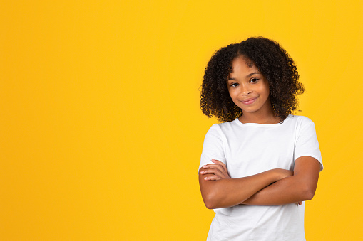 Smiling confident adolescent curly girl in white t-shirt with crossed arms on chest, isolated yellow studio background. Pupil emotions, knowledge, study and education in school, ad and offer