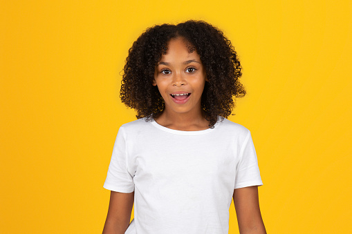 Happy shocked adolescent black girl in white t-shirt with open mouth, isolated on orange background, studio. Surprise reaction, sale, human emotions, good news, study and modern school education