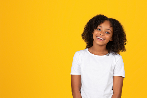 Laughing smiling curly teen african american girl in white t-shirt looking at camera isolated on yellow studio background. Pupil and school education, ad and offer, sale and lifestyle