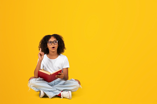 Happy shocked curly teen african american girl in white t-shirt, glasses, reading book, got idea isolated on yellow studio background. Pupil emotions, learning, knowledge at school creat solution