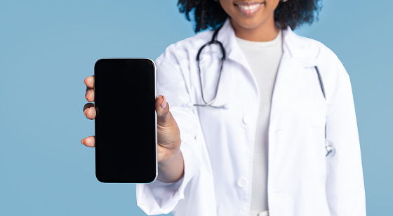 Smiling black millennial lady doctor in white coat show smartphone with empty screen, isolated on blue studio background, cropped. App for work professional recommendation, medicine and health care