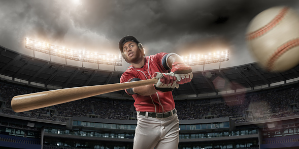 Mid action image of professional baseball player dressed in red and white uniform, swinging bat in floodlit stadium full of spectators under stormy sky