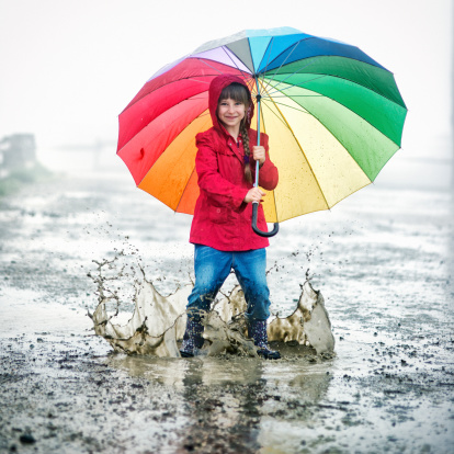Little girl with colorful umbrella having fun jumping in a big puddles during heavy rain. Slightly soft focus.