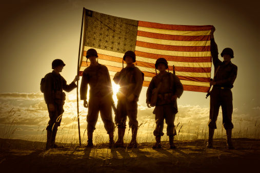 Four WWII Soldiers Holding up a translucent American Flag at sundown.  The sun  behind the flag creates a semi-silhouette of the four war weary soldiers.  Clouds, sun, and sand complete the picture.  All soldiers are in uniform with helmets and rifles, boots etc.