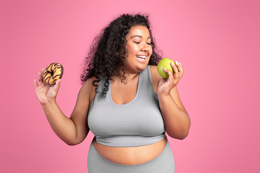Young black body positive lady in sportswear holding donut and biting green apple, choosing healthy food, posing on pink studio background. Proper nutrition, weight loss and diet