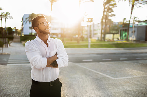 Happy dreamy european businessman in formal wear standing outdoors with folded arms and looking away at empty space. Successful entrepreneur man posing outside