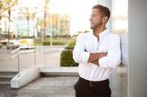Serious confident businessman in shirt posing with crossed arms, looking aside at free space, standing near office building, outdoors. Entrepreneurship and startup