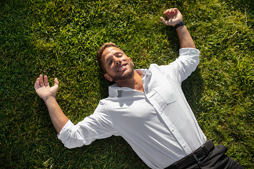 Happy relaxed young businessman in shirt lying on green grass on lawn and resting in park with closed eyes, taking break from work outdoors, above view
